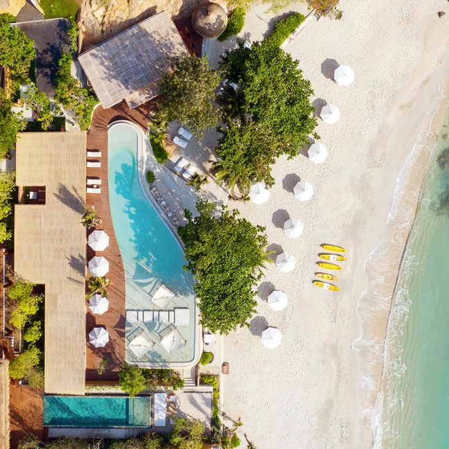 Overlooking a serene beachside setting with a freshwater pool, thatched-roof villas, shade umbrellas, kayaks, and clear blue sea—harmoniously integrated with nature.