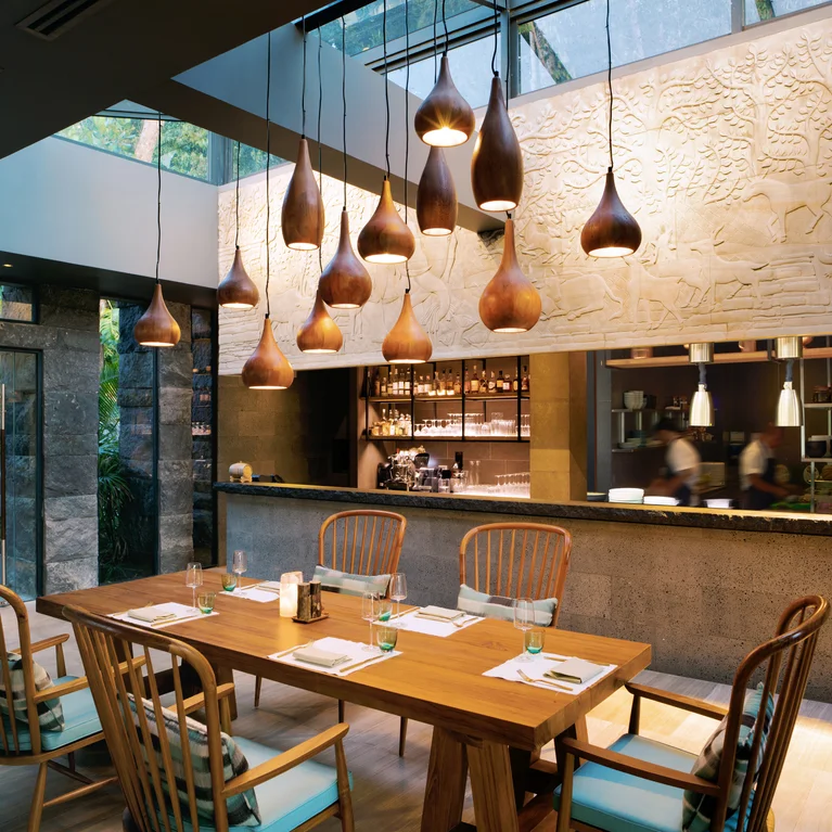 مطعم به طاولات وكراسي خشبية، وإضاءة معلّقة، ومنضدة مطبخ مفتوحة، ونوافذ كبيرة للضوء الطبيعي، وقطعة فنية معروضة على حائط مزخرف.