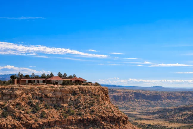 Sustainable retreats set at the cliff’s edge offer expansive views across sunlit terrain beneath a blue sky with gentle clouds.