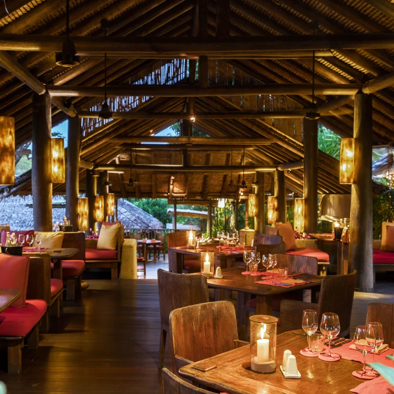 Salle à manger avec mobilier en bois apparent, coussins rouges, éclairage doux, toit de chaume et côtés ouverts donnant sur la végétation. Comprend des allées accessibles. ...