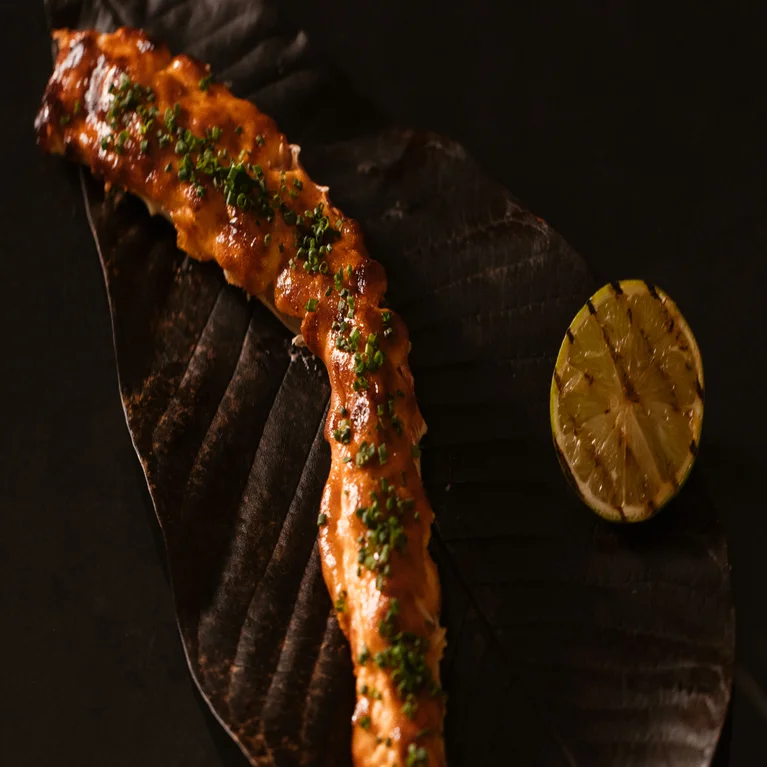 Crab leg with glaze and chives served on a dark leaf plate, accompanied by a grilled lime half.