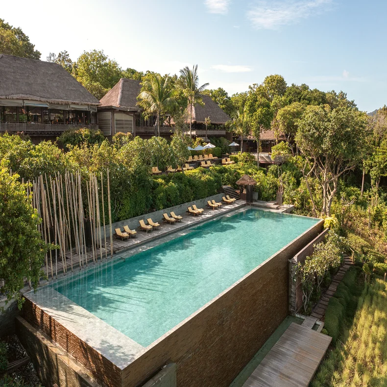 Rectangular infinity pool bordered by lounge chairs and abundant greenery, with timber resort buildings and an open sky, designed for relaxation and mindful well-being.