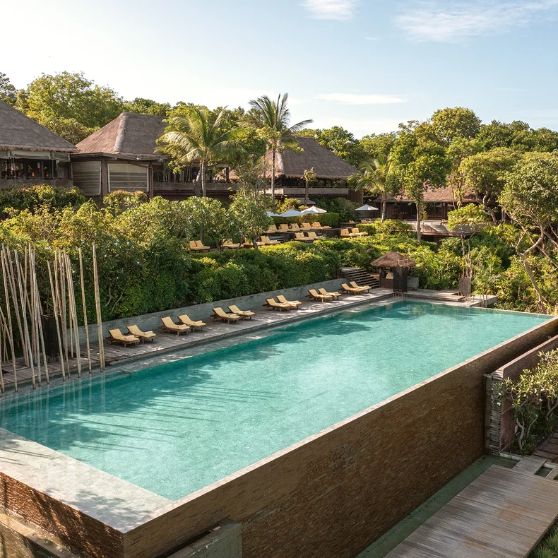 Rectangular infinity pool bordered by lounge chairs and abundant greenery, with timber resort buildings and an open sky, designed for relaxation and mindful well-being.