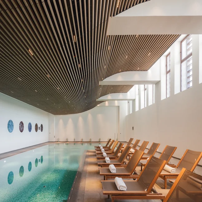 Tranquil water pool area, wooden lounge chairs and soft white towels. Large windows and a slatted ceiling provide daylight for a relaxing lounging experience.