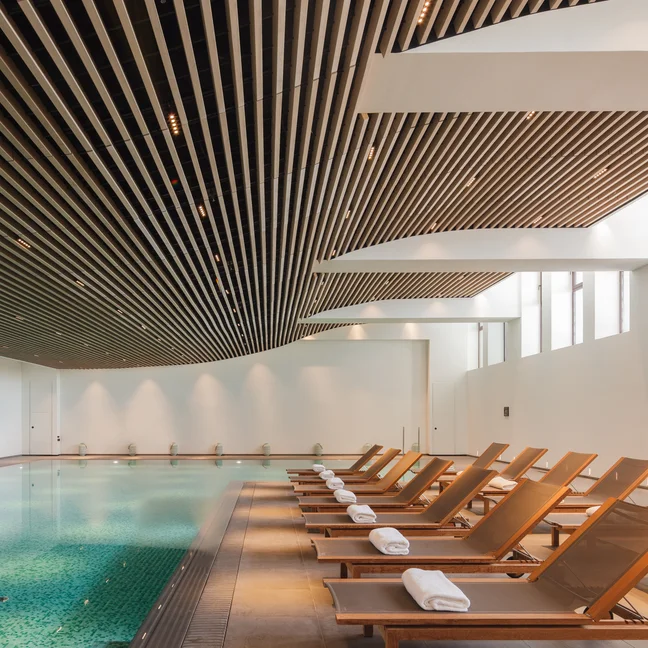 Tranquil water pool area, wooden lounge chairs and soft white towels. Large windows and a slatted ceiling provide daylight for a relaxing lounging experience.