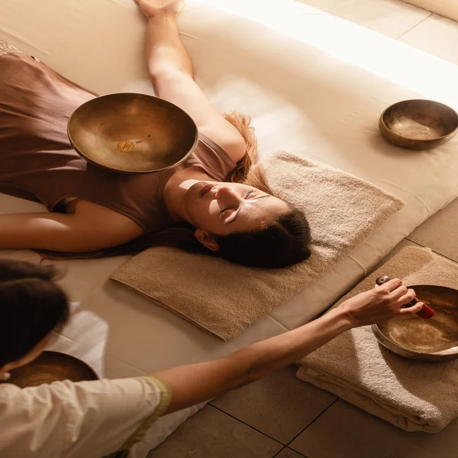 A guest receives sound therapy as singing bowls are placed on the body, with a practitioner facilitating the session in a calming treatment space.