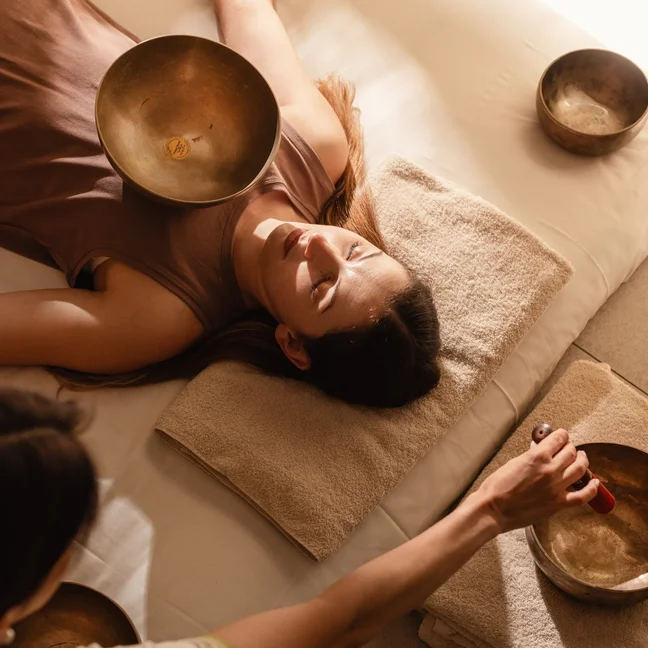 A guest receives sound therapy as singing bowls are placed on the body, with a practitioner facilitating the session in a calming treatment space.