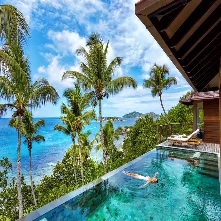 A guest enjoys a tranquil pool with views of palm trees, vibrant greenery, and the blue ocean under a sky dotted with clouds.