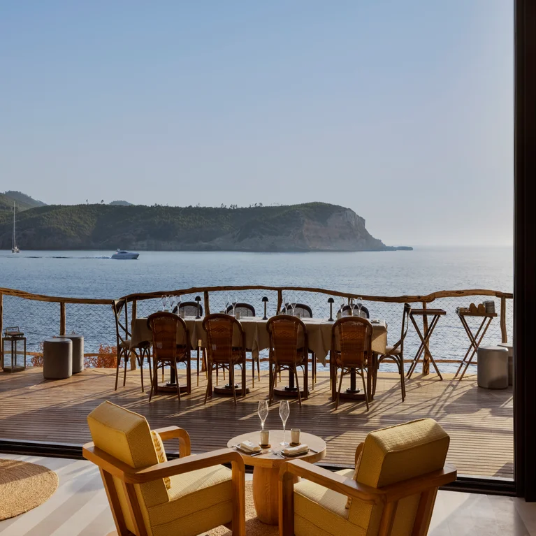 Wooden chairs and tables set on a terrace face the sea and distant hills, visible from a lounge with two yellow armchairs and refreshments. Sunlight fills the serene space.