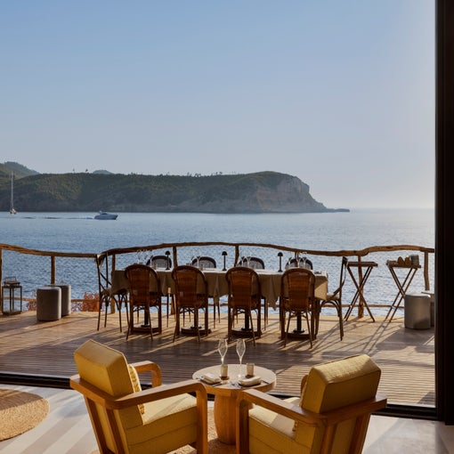 Wooden chairs and tables set on a terrace face the sea and distant hills, visible from a lounge with two yellow armchairs and refreshments. Sunlight fills the serene space.