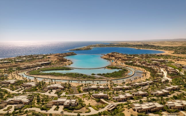 Resort beside calm turquoise lagoons, sandy beaches and low-rise buildings, framed by desert landscape with a clear sky and open sea in view.