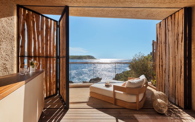A cave suite with tall rough-timber doors thrown wide from a pale plaster interior onto a deck and a low outdoor sofa, the sea glittering straight ahead.