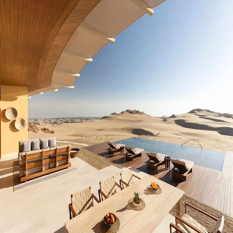 Patio with lounge chairs, dining table, and infinity pool facing desert landscape. Modern design integrates sustainably with the natural environment beneath a clear sky.