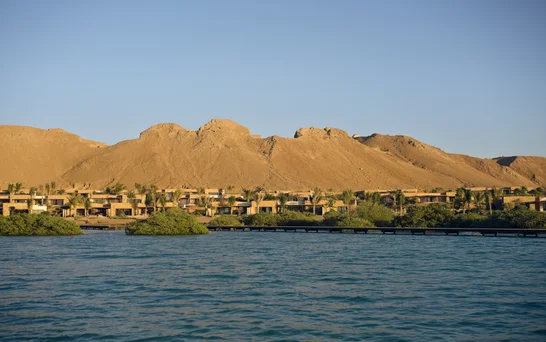 Mountains form a natural backdrop to contemporary buildings and palm trees, with water and green shrubs in the foreground beneath an open sky.