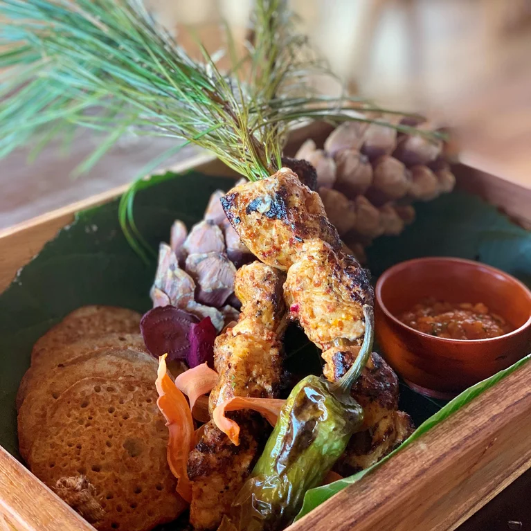 烤肉串、扁面包、烤蔬菜、新鲜香草和一碗酱汁放在香蕉叶上,摆放在一个敞开的木盒中。.