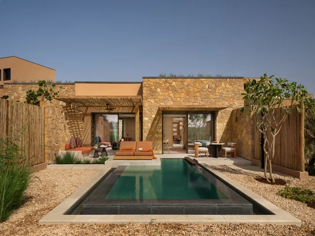 Villa featuring stone walls, a private pool, lounge chairs, shaded outdoor seating areas, and thoughtfully designed native landscaping under clear skies.