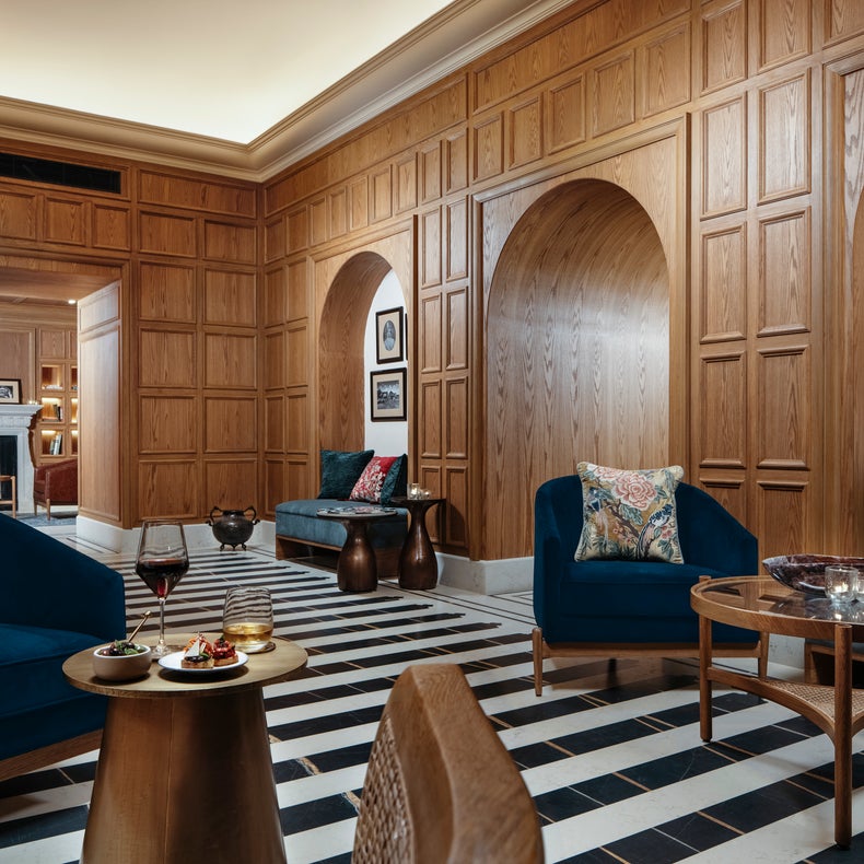 Espacio acogedor con sofás azul oscuro, cojines en tonos tierra, paredes de madera natural, alcobas arqueadas y suelo a rayas. Mesas de relax y cuadros de arte crean un ambiente acogedor.