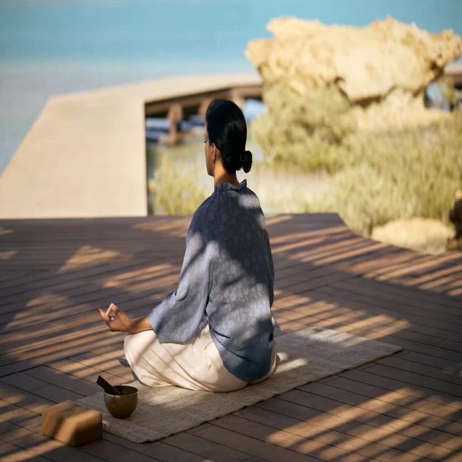 Individual meditating on a mat near calm water, with a singing bowl and block nearby, shaded by trees to blend wellness and nature.