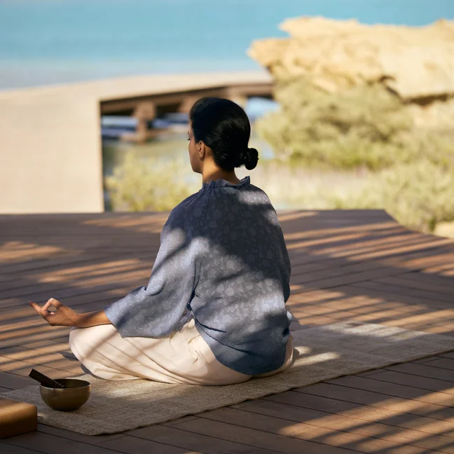 Individual meditating on a mat near calm water, with a singing bowl and block nearby, shaded by trees to blend wellness and nature.