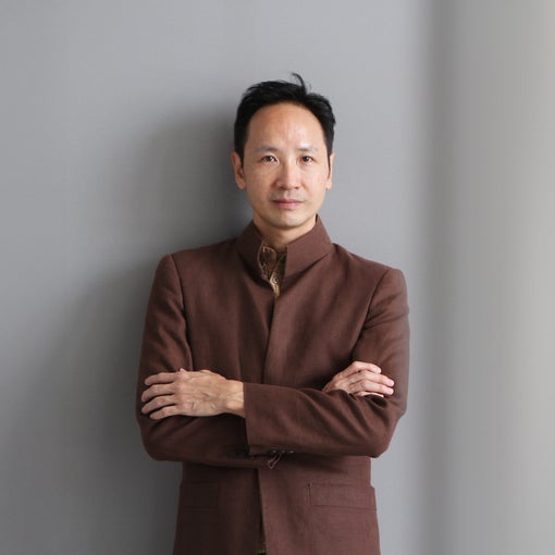 Person wearing a brown suit jacket with arms gently crossed, looking toward the camera, set against a simple grey background.