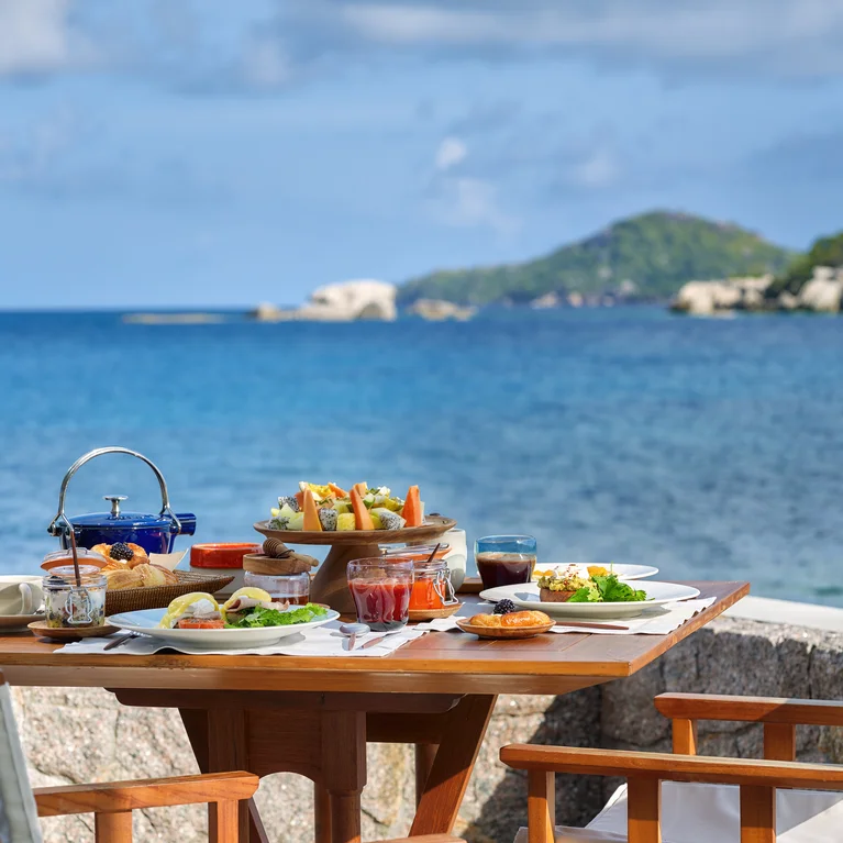 餐桌上摆放着早餐盘和白色椅子,俯瞰着平静蔚蓝的大海、远处的青山和阴霾的天空。.