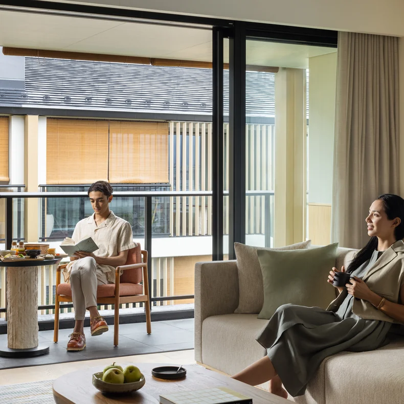A guest enjoys a drink on a sofa in a sunlit living area, while another reads at a balcony table. The space features abundant natural light and open views.