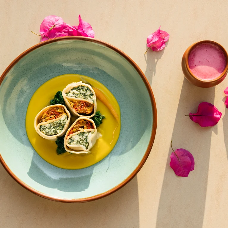 Three filled pasta rolls on yellow sauce, served with bright pink flower petals and a small bowl of pink dip on a beige background.