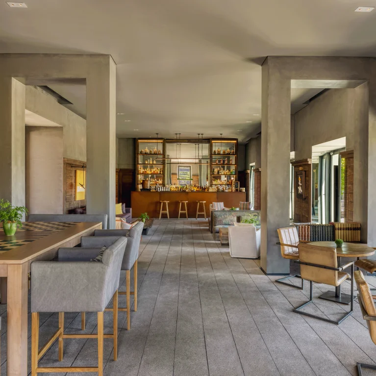 صالة مع ببار وطاولات مرتفعة وكراسي ناعمة بذراعين وتفاصيل من الخشب الطبيعي ونوافذ كبيرة تستقبل ضوء النهار.