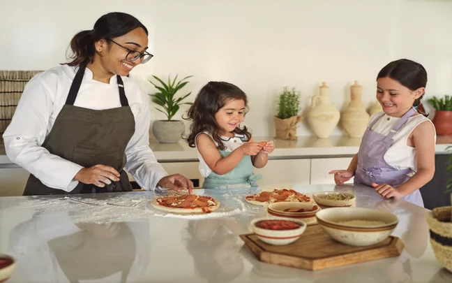 Guests of different ages prepare pizzas together in a sunlit kitchen, using fresh ingredients arranged in bowls on the counter.
