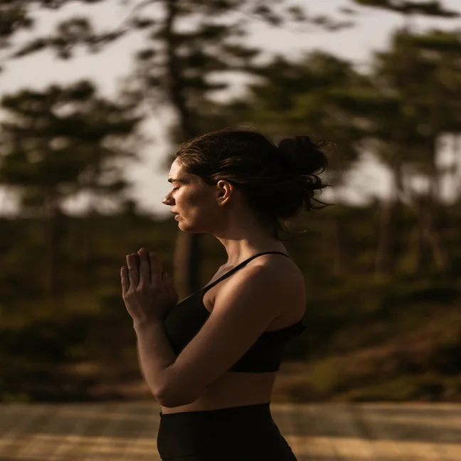 A person practices yoga outdoors, eyes closed and hands together at chest level, dressed in activewear and surrounded by lush greenery.
