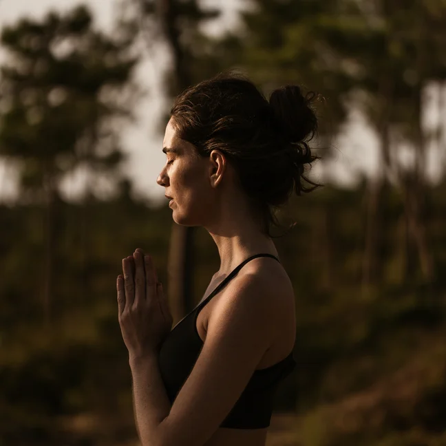 A person practices yoga outdoors, eyes closed and hands together at chest level, dressed in activewear and surrounded by lush greenery.