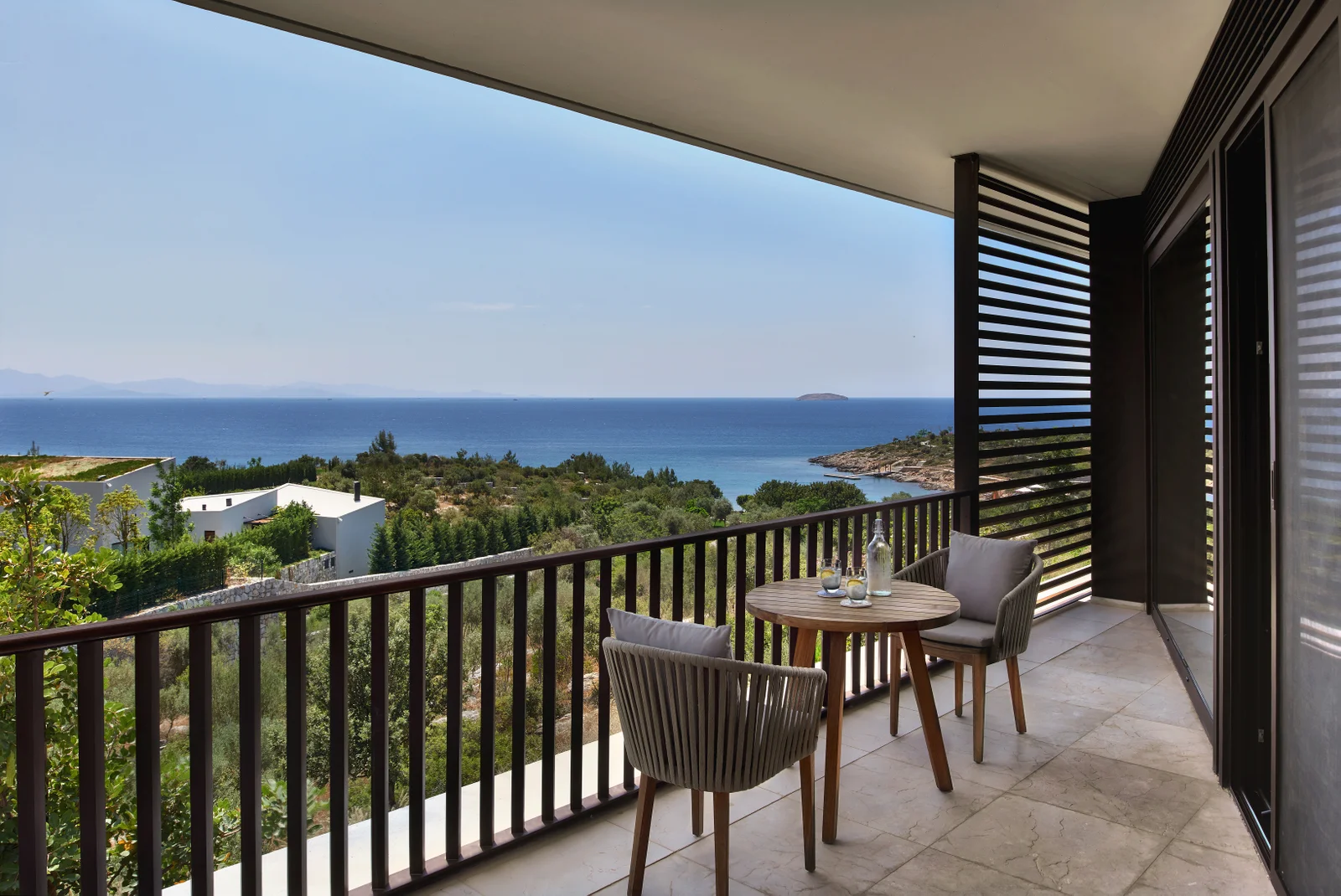 A junior suite balcony with two woven chairs and a round timber table, a louvered screen at one end and the bay and an island beyond.