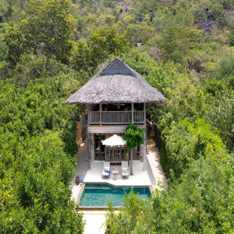 Sustainable villa with thatched roof, outdoor pool, sun loungers and a shade umbrella, set among abundant tropical plants.