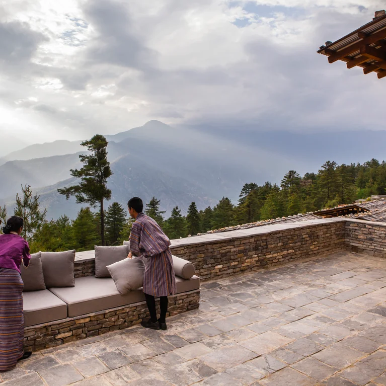 Team members set up cushions on a stone patio with views of forested mountains, while soft sunlight filters through clouds overhead.