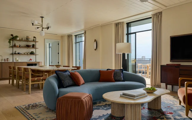 Spacious living and dining space featuring a blue curved sofa with orange cushions, round tables, large windows, wooden dining table and shelves displaying decor on light walls.