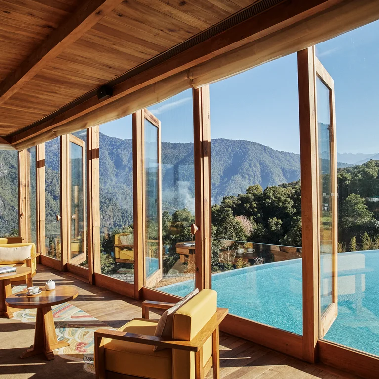 Lounge featuring yellow chairs and wooden details faces a mountainside infinity pool, with forested hills visible through large glass windows.