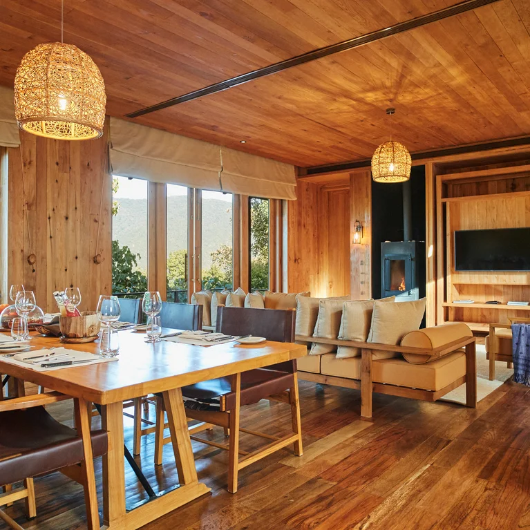Dining and living area with natural wood walls, floor, and ceiling; large windows bring in light. Includes dining table, sofas, TV, and gentle ambient lighting.
