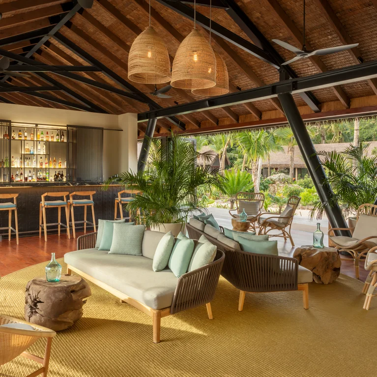Lounge area with natural fiber seating, low wooden tables, and abundant greenery; an inviting bar with stools sits beneath a timber ceiling, opening to the outdoors.