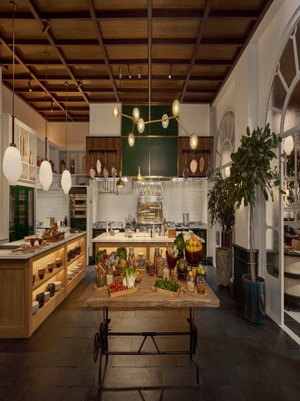 Kitchen features a natural wood ceiling, suspended lighting, accessible open shelving, and a central table displaying an assortment of fresh produce and herbs.