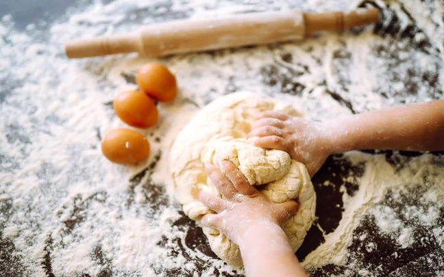 Hands gently prepare dough on a lightly floured surface, with a trio of eggs and a rolling pin set in the background, evoking mindful culinary practice.