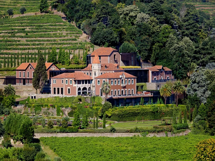 Villa with red-tiled roofs set among vineyards, mature trees, and structured gardens on a hillside, highlighting natural surroundings and sustainable landscaping.