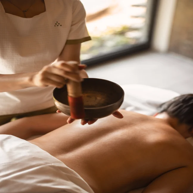 A practitioner uses a singing bowl above a guest lying on a massage table during a wellness treatment.