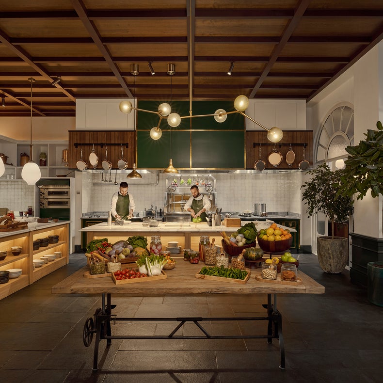 Chefs prepare dishes in a bright kitchen featuring sustainable wood accents, globe pendant lighting, and a table with fresh produce and glass jars.