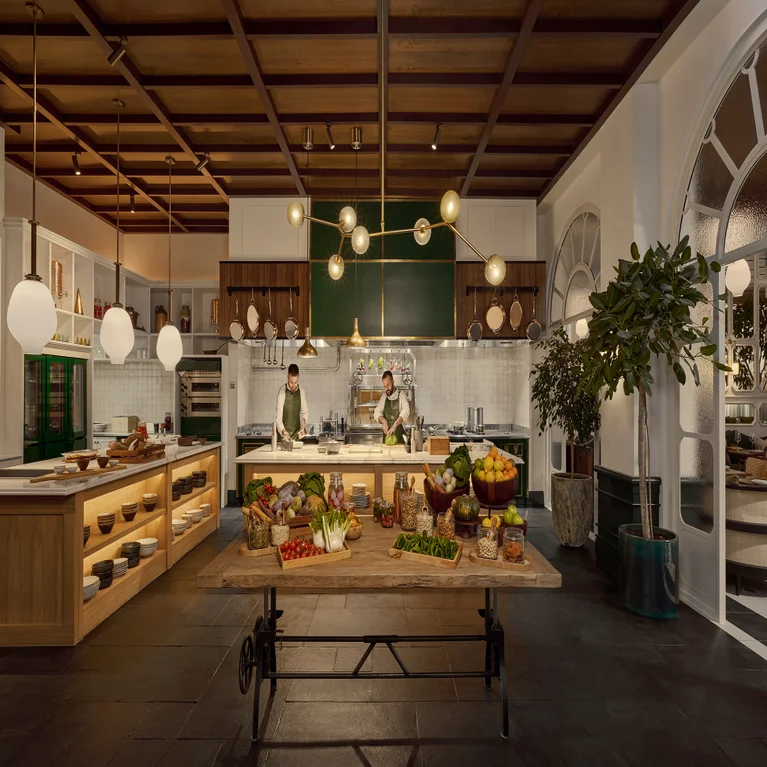 Chefs prepare dishes in a bright kitchen featuring sustainable wood accents, globe pendant lighting, and a table with fresh produce and glass jars.