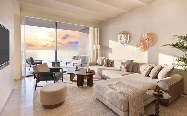 Living space featuring natural materials, comfortable seating, and expansive window leading to balcony with sea and sunset views, designed for wellbeing and relaxation.