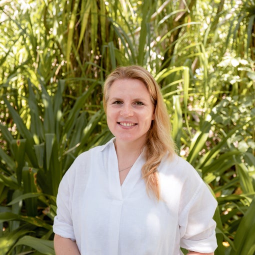 A guest with light hair and a white shirt stands outdoors, smiling near vibrant green plants, reflecting Six Senses’ focus on nature and wellbeing.