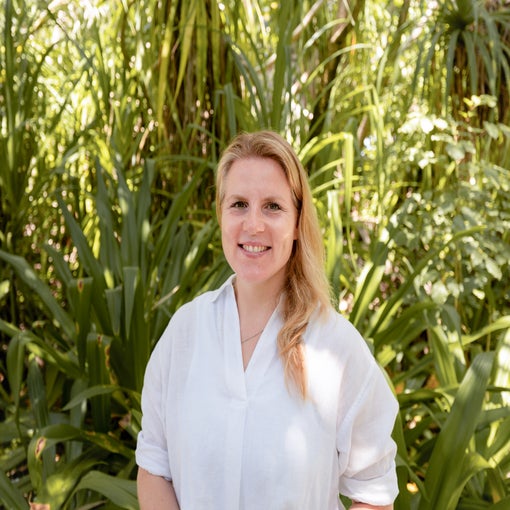A guest with light hair and a white shirt stands outdoors, smiling near vibrant green plants, reflecting Six Senses’ focus on nature and wellbeing.