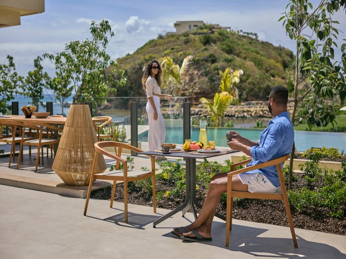 A guest enjoys an outdoor meal while another walks by the pool, surrounded by natural views and gentle hills in the background.