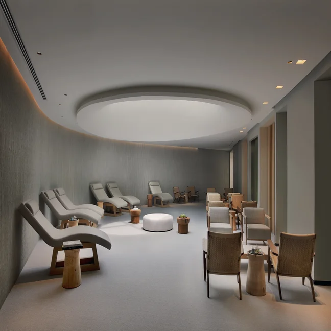 Curved-walled lounge with reclined chairs on one side, wicker seating with small tables opposite, and gentle light from a circular ceiling fixture.