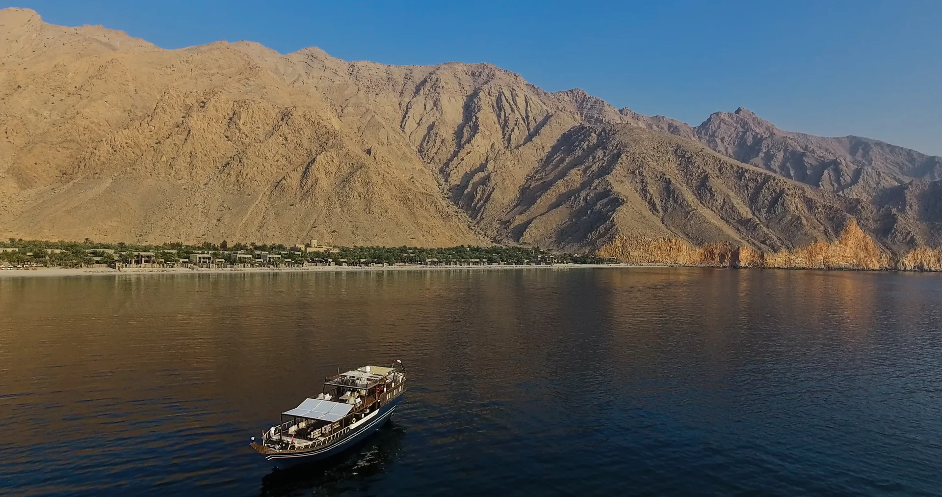 The resort seen from the water as a thin green band of palms along the shoreline, dwarfed by the bare ochre ridges of the Musandam peninsula, with a traditional wooden dhow anchored in the foreground.